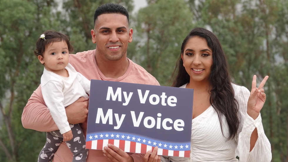 A young Hispanic family smiles for the camera while holding up a sign saying "My Vote My Voice".