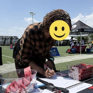 A Black female wearing glasses and a brown and black shirt leans over a table signing a piece of paper at a community event.