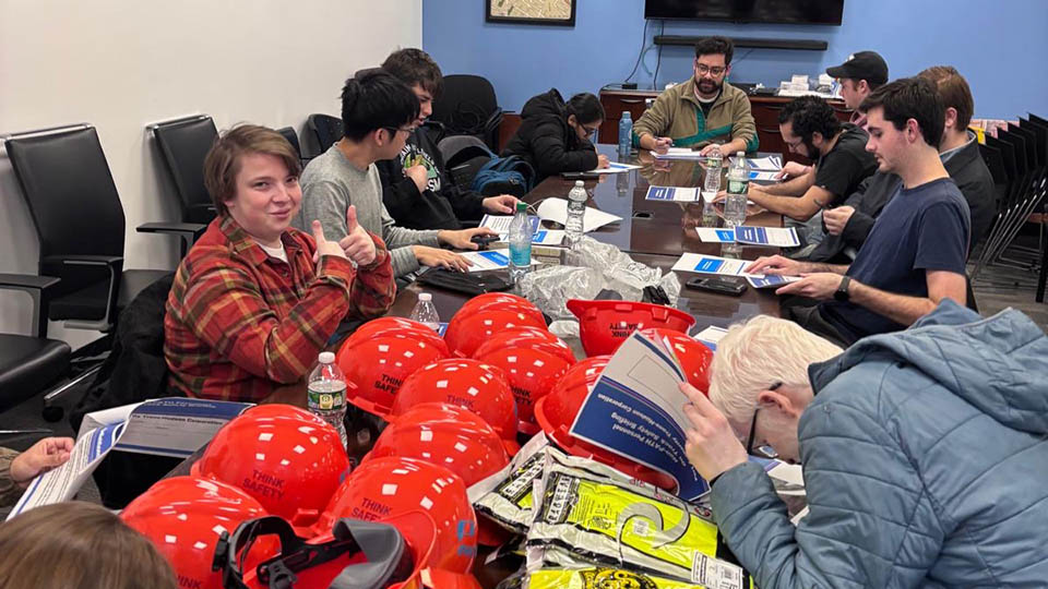 Students sitting at a conference table with orange hard hats in the foreground participating with Bloustein Enthusiasts and Advocates for Transportation (BEAT) took part in an exclusive after-hours PATH (Port Authority Trans-Hudson) tour.