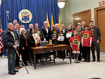 A large group of individuals, including members of the public, legislators, and New Jersey Gov. Phil Murphy, at the New Jersey Statehouse in Trenton, NJ to celebrate the signing of the Target Zero Commission Law. Individuals are standing in large room with a blue background around a dark brown table.