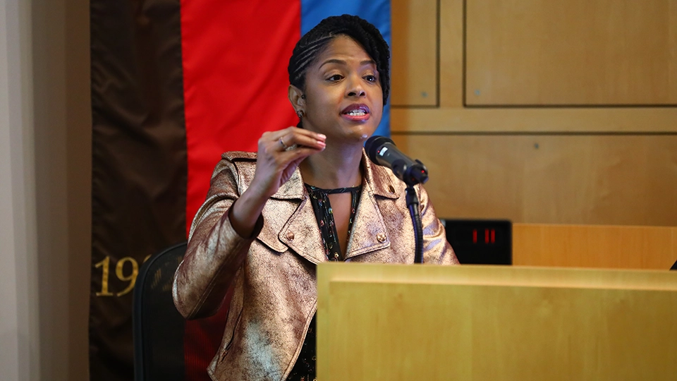 Dr. Chris T. Pernell, an African-American woman, stands behind a podium speaking into a microphone, mid-gesture with one hand raised as if emphasizing a point. She wears a metallic gold jacket over a dark patterned dress. Behind her, a vertical banner with bold red, blue, and black stripes adds color to the indoor lecture setting.