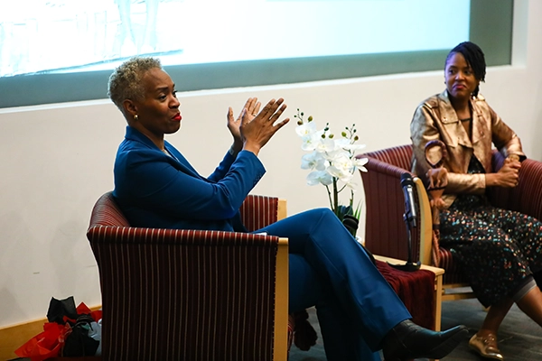 Two women sit on a stage in upholstered armchairs during a panel discussion. The woman on the left, Dr. Patti O'Brien Richardson, is an Associate Teaching Professor | Bloustein School Chief Community and Belonging Officer. She wears a bright blue suit, leans slightly forward and gestures with both hands while speaking. The woman on the right is the lecture speaker, Dr. Chris T. Pernell, director of the NAACP Center for Health Equity. Dr. Pernell wears a gold jacket and patterned dress, sits back and looks toward Dr. O'Brien Richarson. A small table between them holds a microphone and a vase of white flowers, with a projection screen in the background.