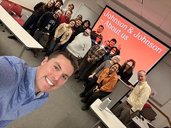 Trevor Fuller, Senior Manager, Transportation Operations is in the the foreground, smiling and taking a selfie. A group of about 15–20 smiling, relaxed students and guests stand behind him in a classroom. They are in front of presentation slide on a screen reads “Johnson & Johnson – About us.” 