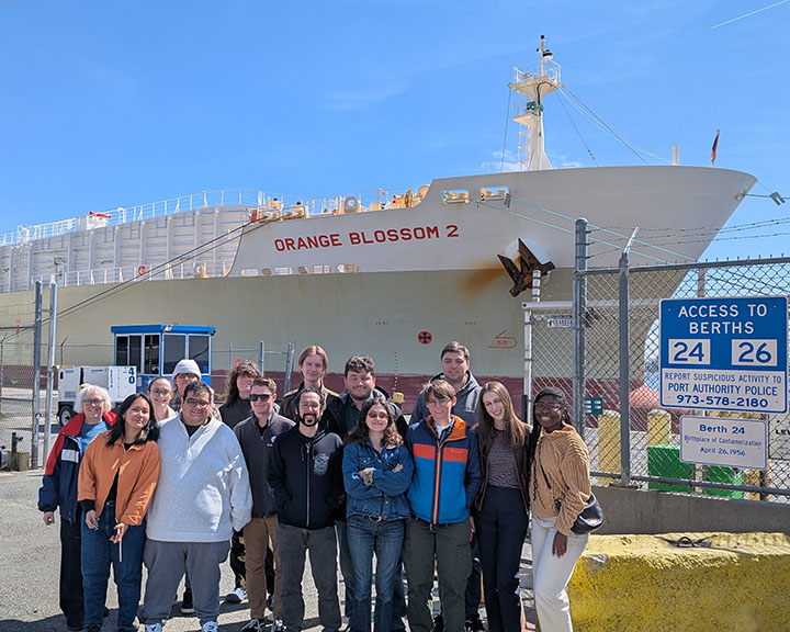 A diverse group of students from the Bloustein School's Freights & Ports master's level class visit Berth 24 at Maher Terminal at Port Newark/Elizabeth on a bright sunny. They stand in front of a white tanker ship with Orange Blossom 2 on the side, a ship dedicated to bring orange juice into the region for processing at a nearby facility. Signs indicating the berth location of the ship are posted on a gate to the students' right side.