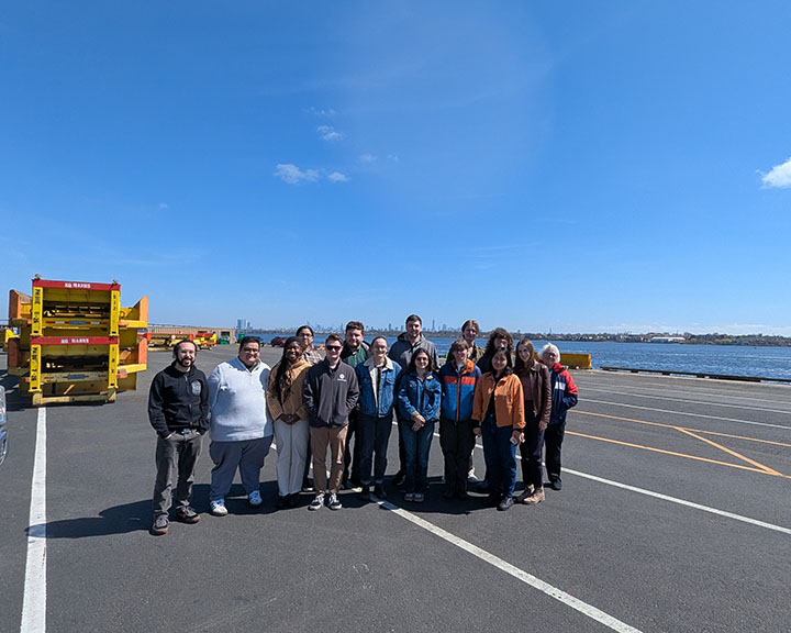 A diverse group of students from the Bloustein School's Freights & Ports master's level class visit Maher Terminal at Port Newark/Elizabeth on a bright sunny day. They are on a road in front of water. A yellow rubber-tired Gantry (RTG) crane is behind them.