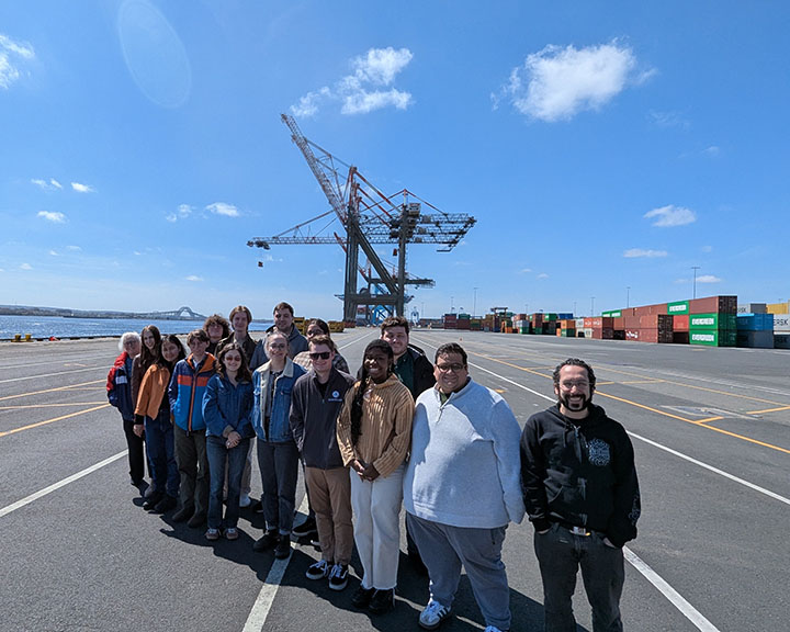 A diverse group of students from the Bloustein School's Freights & Ports master's level class visit Maher Terminal at Port Newark/Elizabeth on a bright sunny day. They are on a road in front o water. A ship-to-shore (STS) container crane and multicolor shipping containers are in the background.